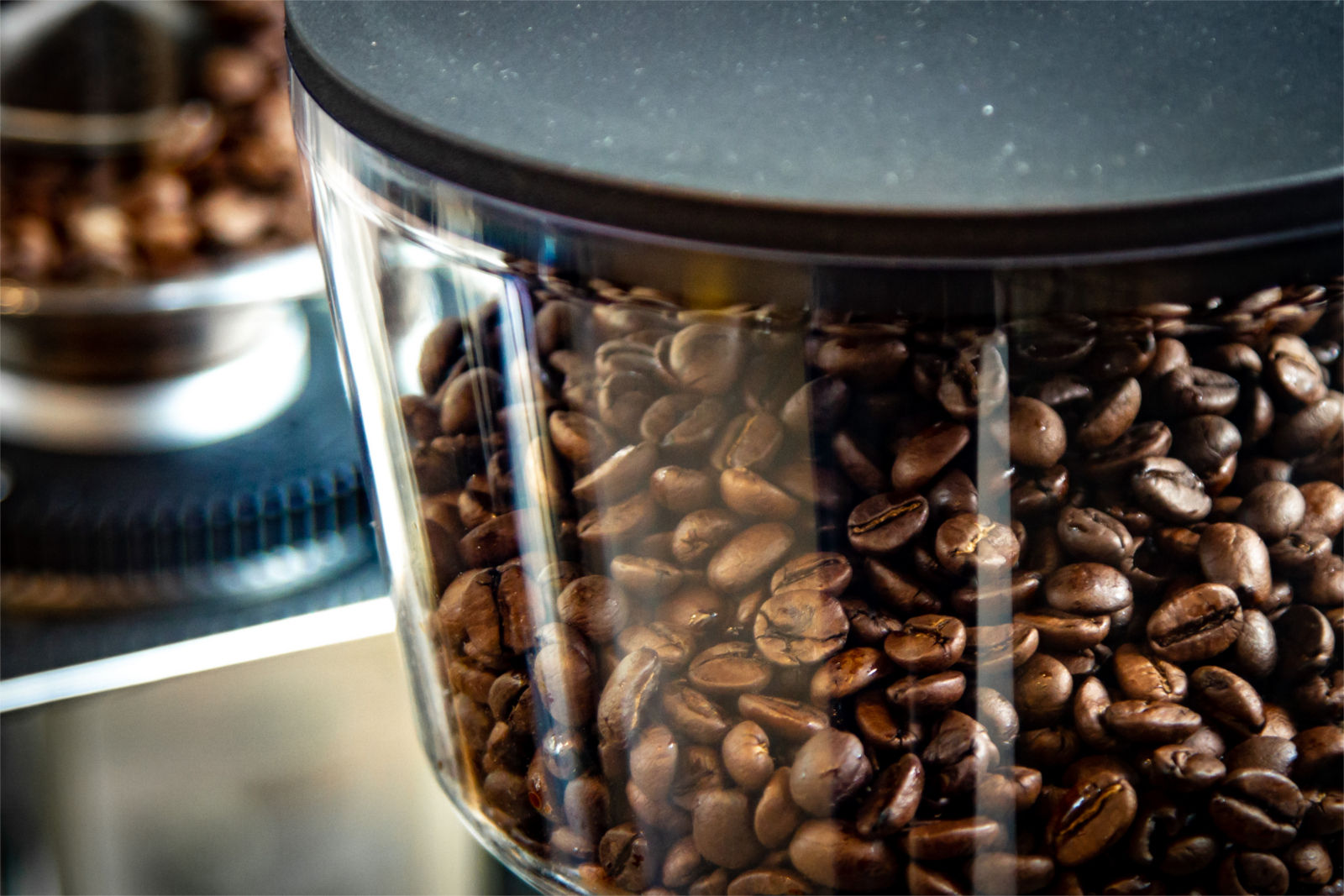 Ein durchsichtiger Behälter mit dunklen Kaffeebohnen und einem schwarzen Deckel steht im Vordergrund, während im Hintergrund ein Kaffeemühlenbehälter zu sehen ist.
